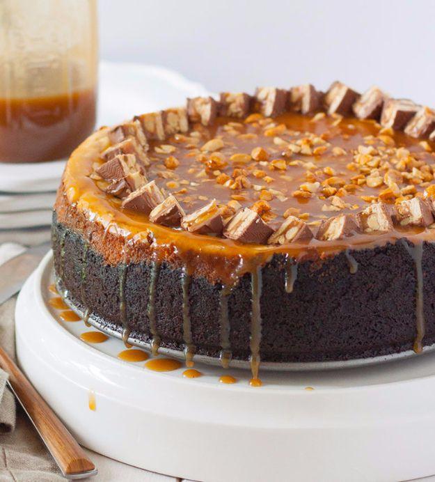 Snickers cheesecake on a white stand with caramel sauce dripping down the sides over a dark Oreo crust