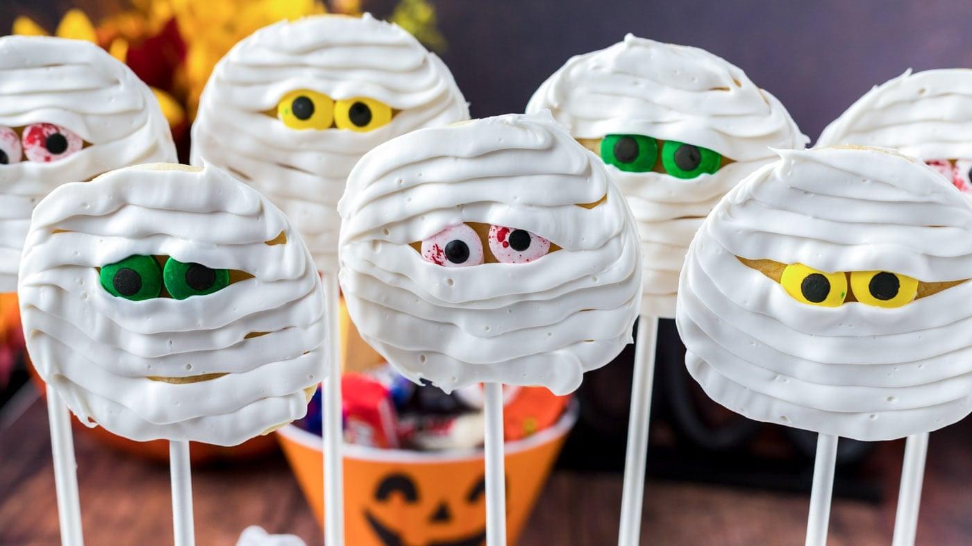 Round mummy cookie pops wrapped in white icing bandages with candy eyes and lollipop sticks for Halloween
