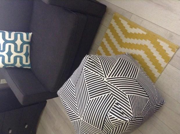 Black and white geometric print cube floor pouf beside a grey sofa and a yellow chevron rug in a living room