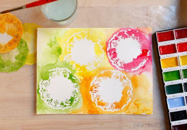 Watercolor painted through paper doilies in yellow, green, pink and orange on paper beside a paint palette