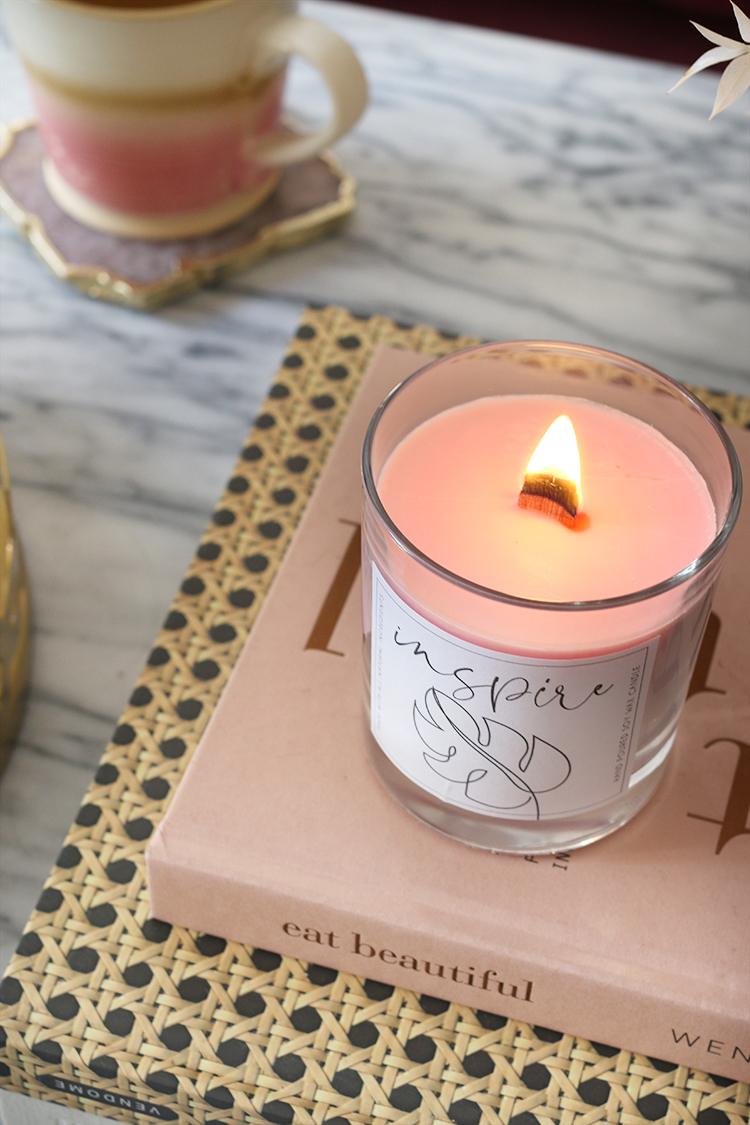 Lit pale pink soy candle with a wood wick in a clear glass holder sitting on stacked books and a rattan tray