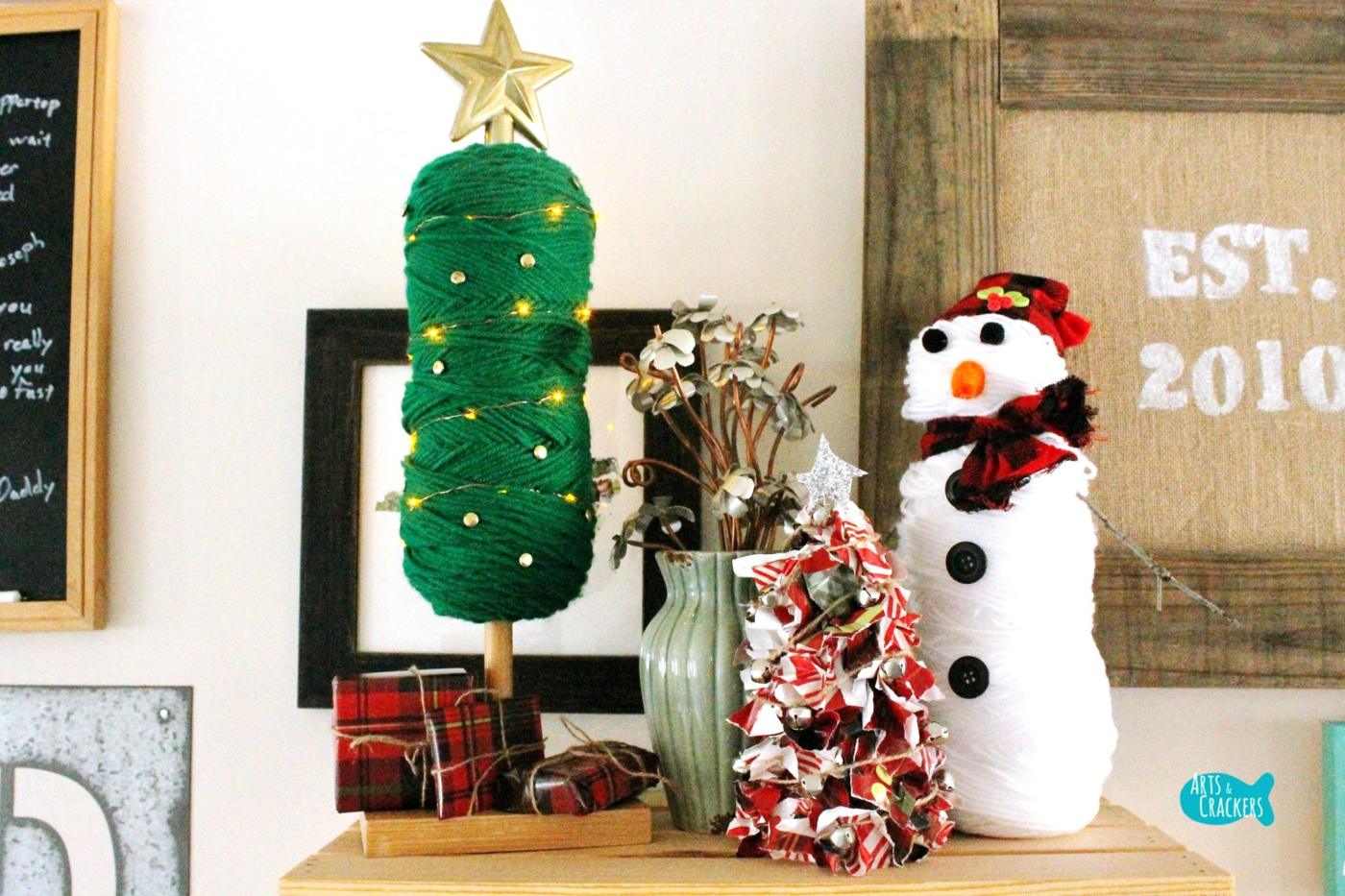A green yarn skein tree on a wooden post wrapped in fairy lights with a gold star, beside a rag tree and a yarn snowman