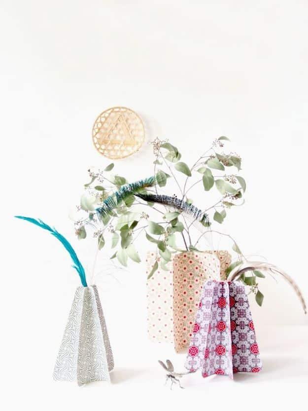Four vases wrapped in folded patterned paper covers in gray, cream and pink, holding eucalyptus and a teal feather