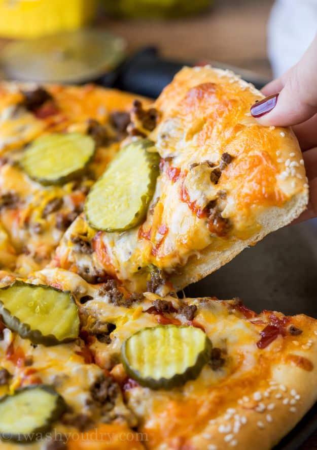 Hand lifting a cheeseburger pizza slice topped with beef, cheddar and dill pickle slices from a sesame crust