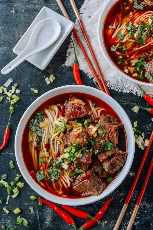 Spicy beef noodle soup in a white bowl of red broth with beef chunks, noodles and scallions, red chilies beside
