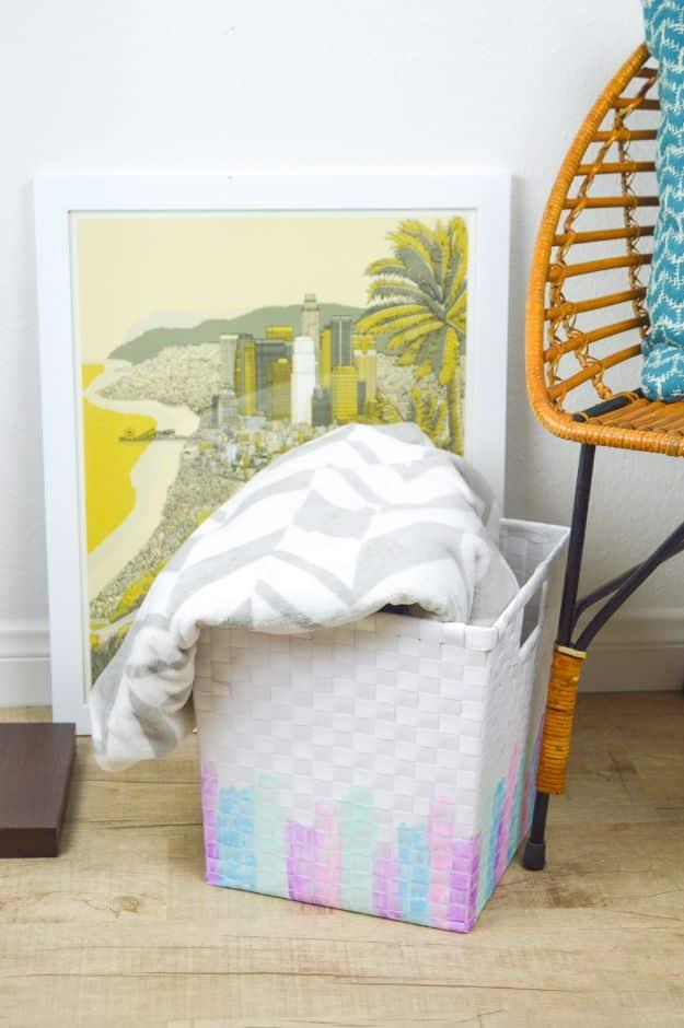 White woven basket painted with pink, mint and yellow watercolor style brush strokes, holding blankets by a chair