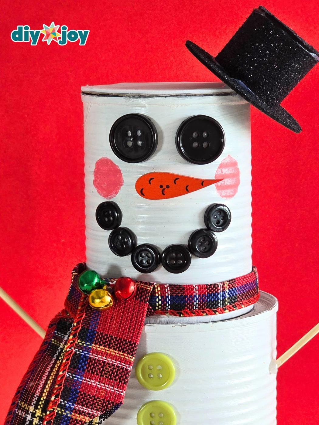 Tin can snowman with button eyes and smile, an orange carrot nose, a plaid ribbon scarf with bells and a glitter foam top hat