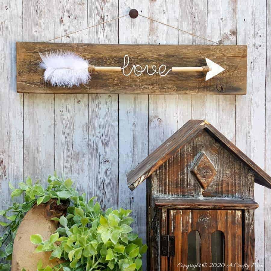 Wire word love on a wooden arrow with a white feather and soda can arrowhead, mounted on a weathered wood plank