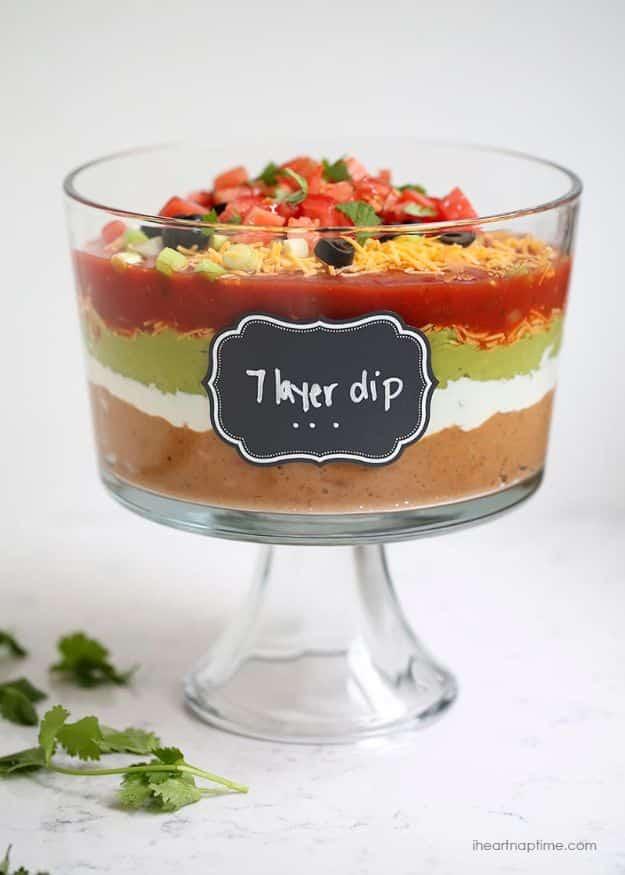 Seven layer dip in a glass dish showing beans, sour cream, guacamole, salsa, cheese, tomatoes and olives