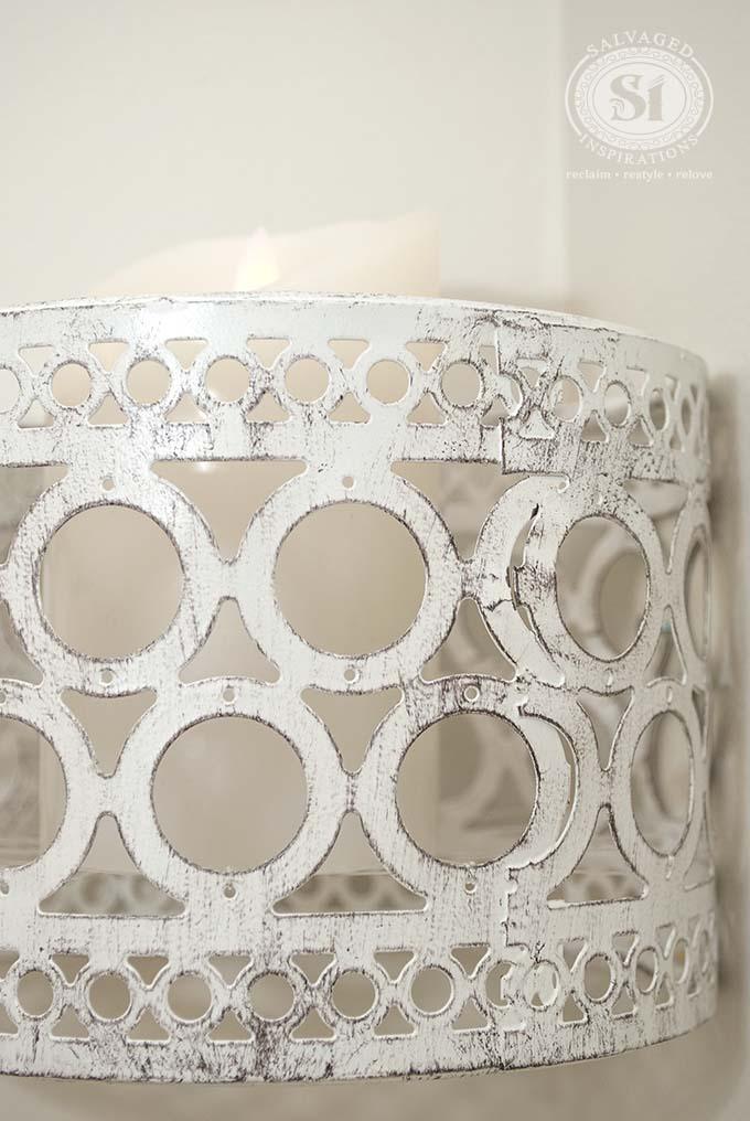 Distressed metal candle shade, a white distressed pierced metal cylinder lit from within by a candle on a wall