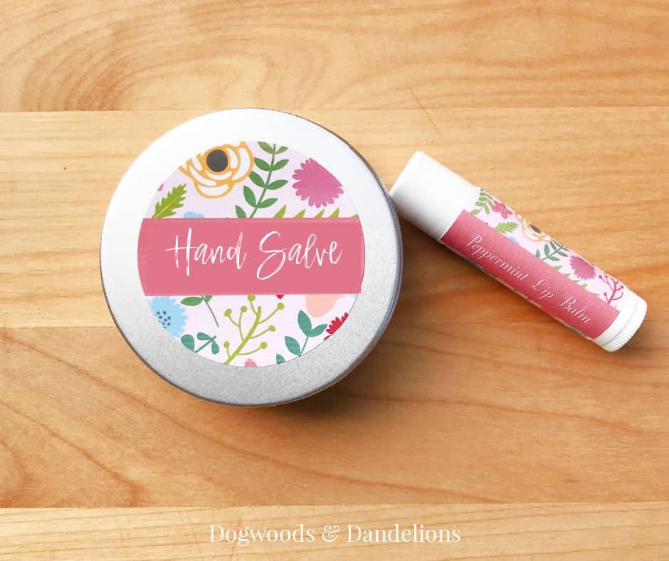 Small round metal tin of hand salve with a floral hand salve label beside a matching lip balm tube on a wood table