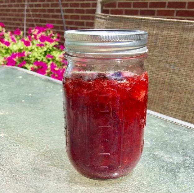 Homemade strawberry jam in a glass mason jar sitting on a patio table in front of a brick wall and flowers