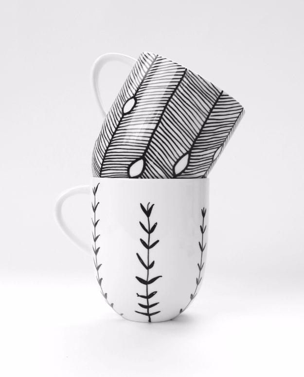 Hand painted porcelain mugs, two white mugs drawn with fine black feather and leaf patterns on a pale background