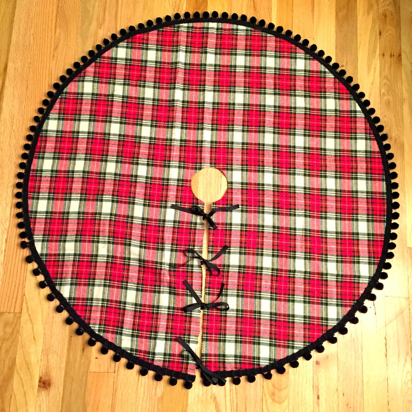 Round red and white plaid Christmas tree skirt edged with black pom pom trim laid out on a wood floor
