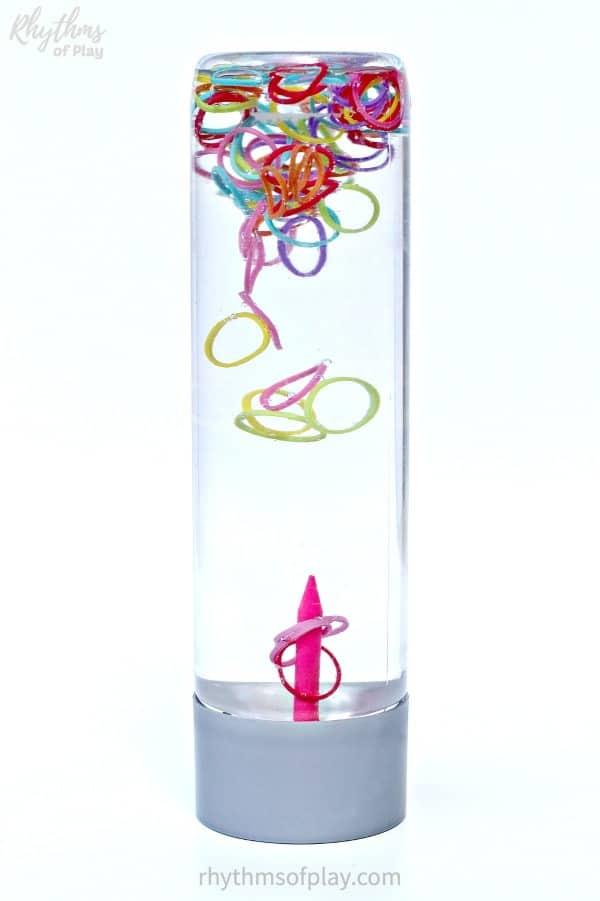 A clear bottle sensory toy with colorful loom band rings floating in water above an upright pink pencil peg in the base