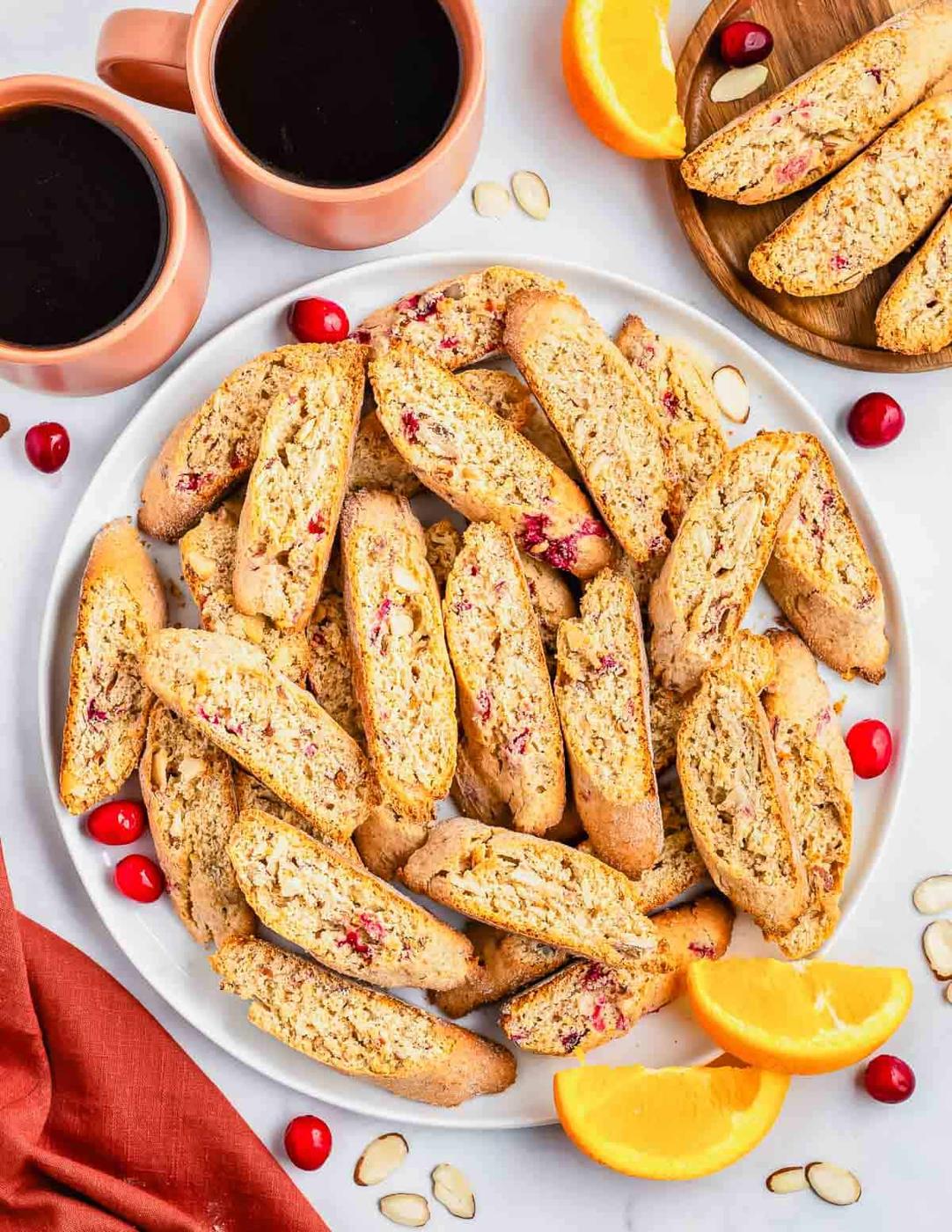 A platter of cranberry almond biscotti with fresh cranberries and sliced almonds beside two mugs of coffee and orange slices