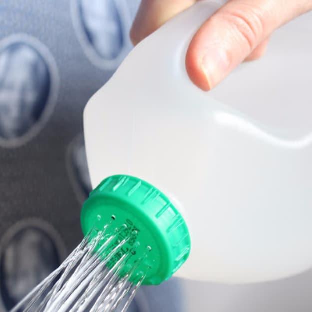 Hand tipping a plastic milk jug with a green perforated lid, water spraying out in fine streams over a seed tray