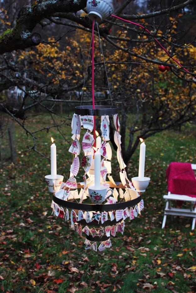 Outdoor mosaic chandelier hung from a bare tree branch with lit white candles, autumn leaves and a red draped table behind