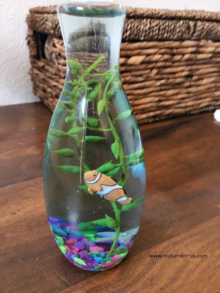 A small plastic bottle aquarium with purple and green gravel, a plastic plant and a toy clownfish floating inside on wood