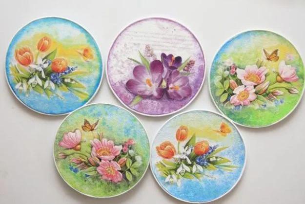 Five decoupaged CDs papered white and decorated with painted floral napkin designs in pink, purple and blue