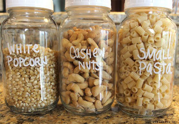 Three large glass jars of white popcorn, cashew nuts and small pasta hand lettered in white on a granite counter