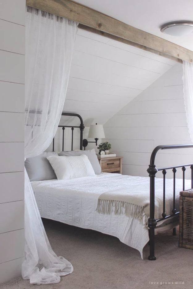 DIY curtain canopy of white lace panels hanging from a stained wood beam over an iron bed in a white shiplap attic bedroom