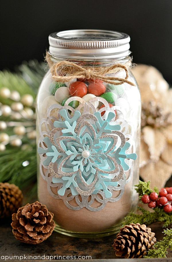 Glass mason jar filled with layered hot cocoa mix and marshmallows, trimmed with a silver doily and paper snowflakes