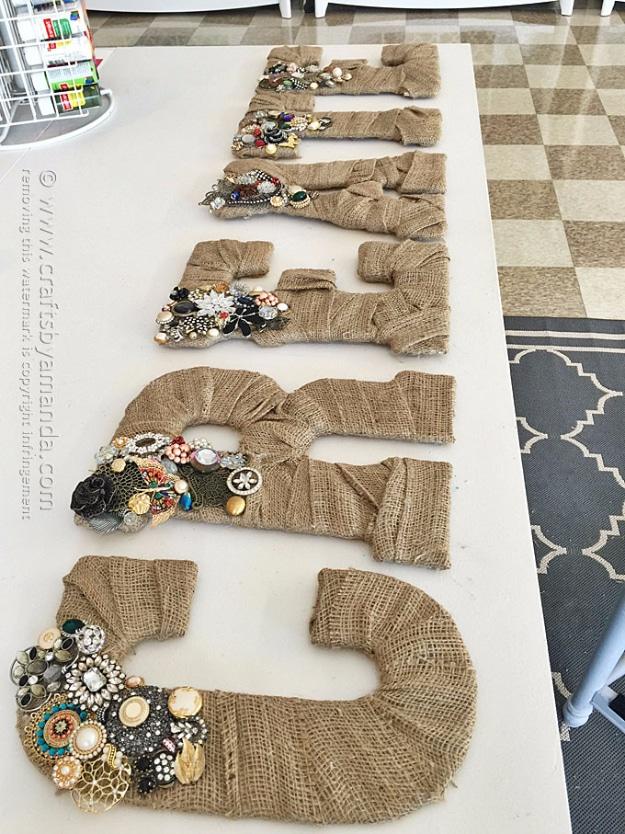 A burlap wrapped letter H trimmed with small rhinestones and a ribbon leans against a white wall on a painted shelf