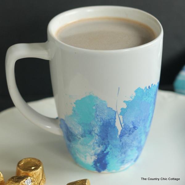 White ceramic mug of coffee with blue and teal marbled paint swirling up from the base, on a white table with candy