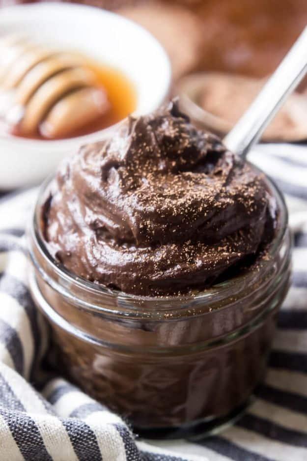 Paleo chocolate avocado pudding in a glass jar dusted with cocoa, a spoon resting in it on a striped cloth