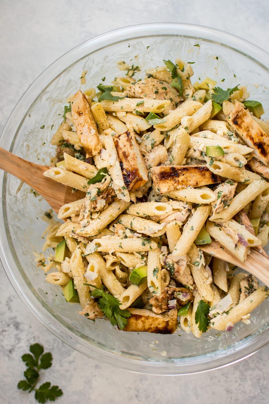 Grilled chicken pasta salad in a large glass bowl with penne, grilled chicken, herbs and a wooden spoon