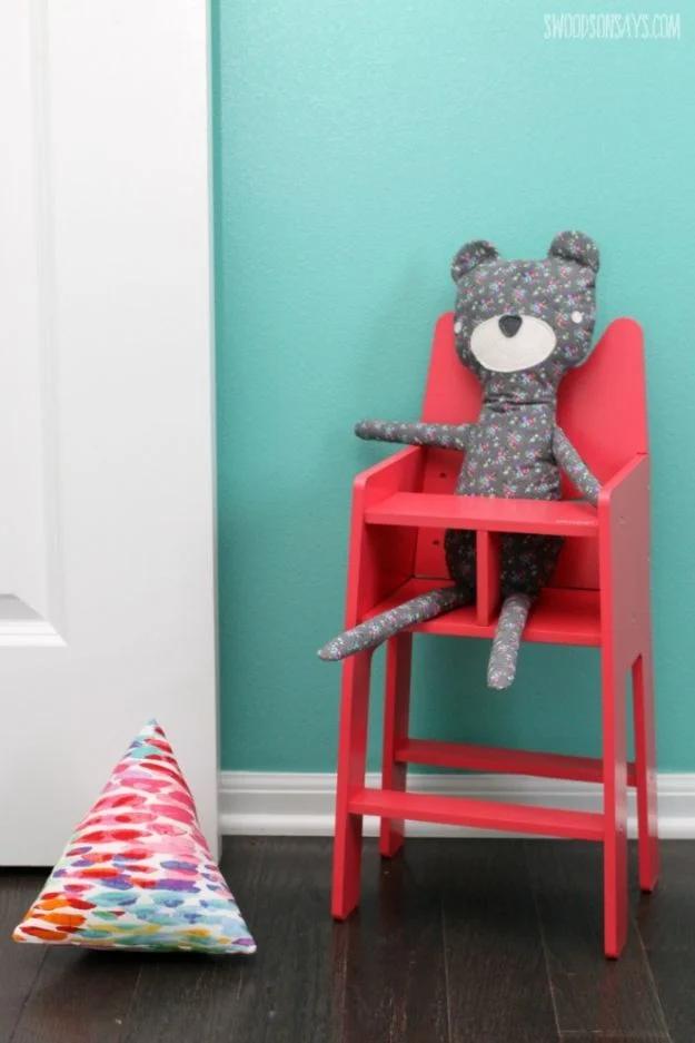 Fabric doorstop sewn in a bright multicolored print sitting on a dark floor beside a red doll high chair holding a grey bear