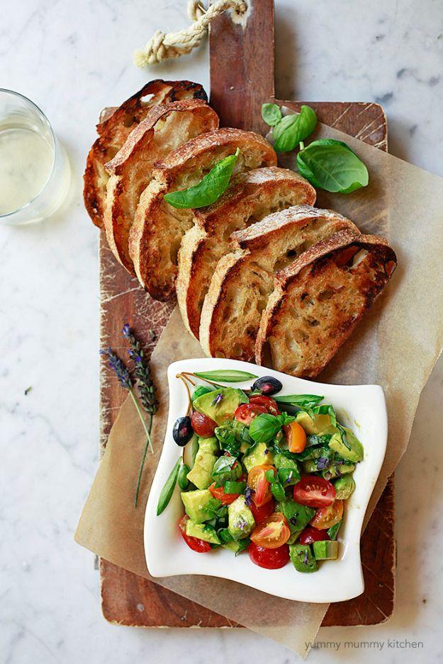 Sliced toasted baguette on a wooden board beside a white bowl of diced avocado, tomato and basil topping