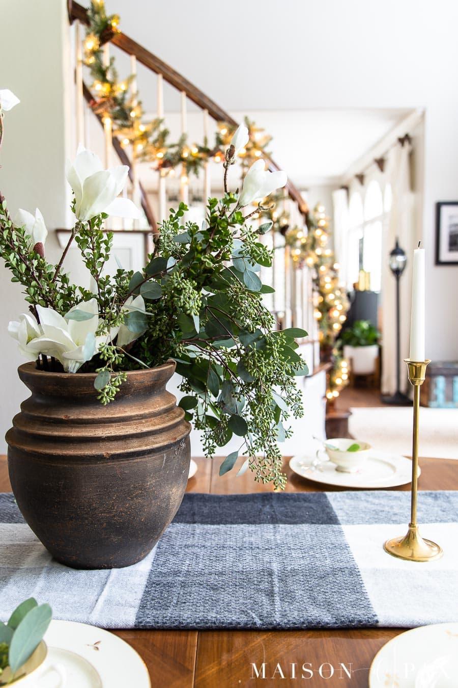 A brown pot of faux magnolia, boxwood and seeded eucalyptus on a black and white plaid runner beside a brass candlestick