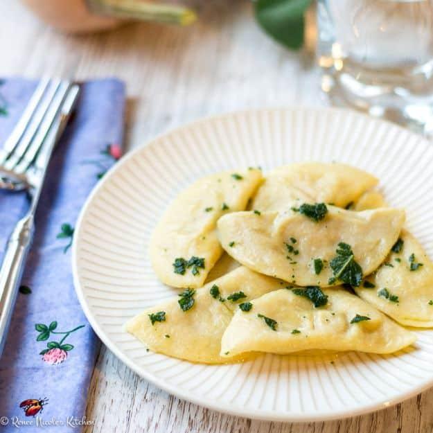 Homemade butternut squash ravioli on a white plate dressed with melted butter and chopped fresh green herbs