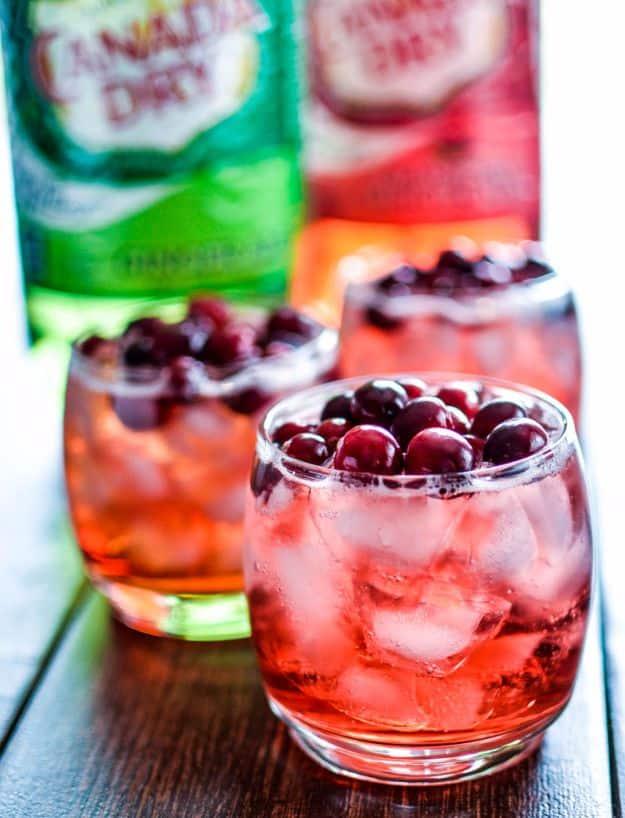 Glasses of cinnamon and cranberry bourbon spritzer over ice topped with fresh cranberries, ginger ale behind