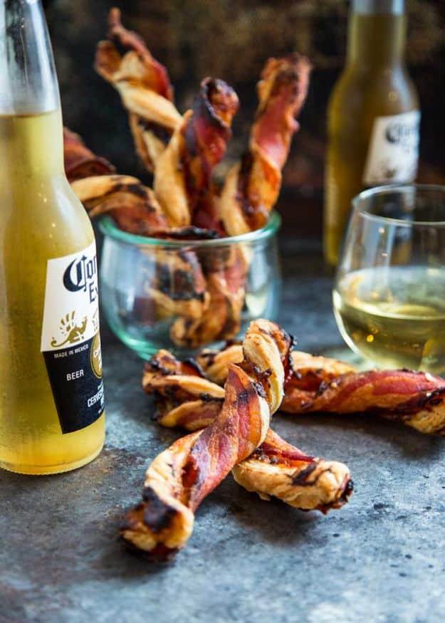 Spiralled bacon and puff pastry cheese twists standing in a glass jar on dark slate, a beer bottle and wine behind