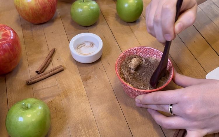 Homemade Bread - two hands stirring cinnamon and sugar together with a spoon in a small red bowl, with whole apples and cinnamon sticks on the wood table