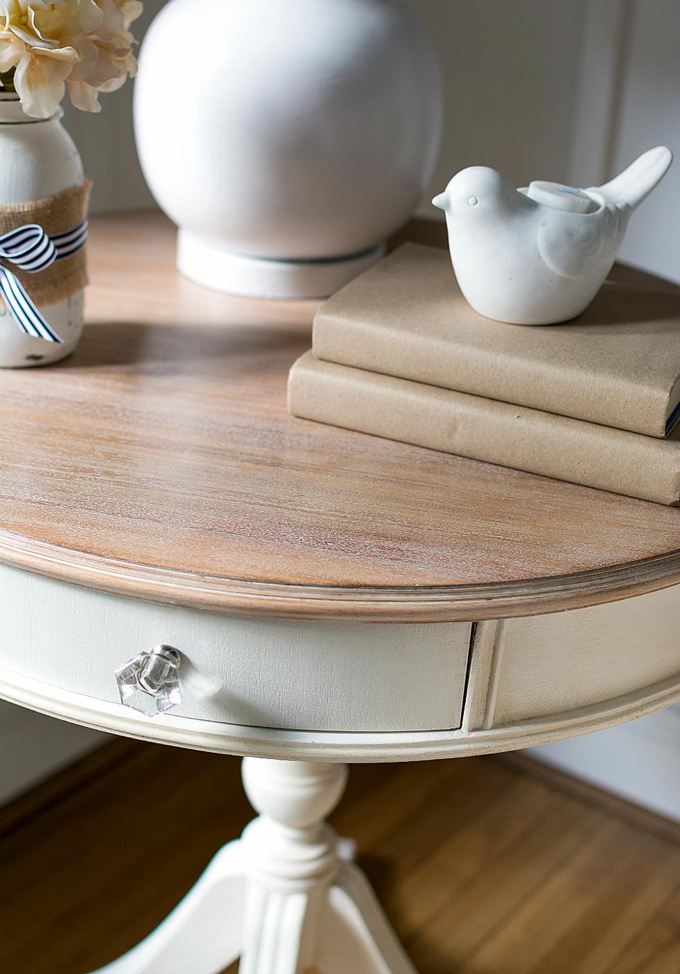 Cream painted round drum table with a soft limewashed wood top, styled with a ceramic bird, stacked books and a vase