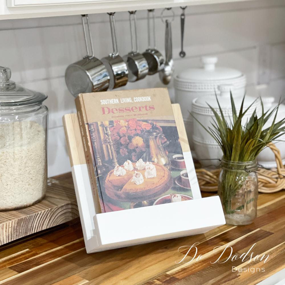 A two toned wood and white cookbook stand holding a vintage dessert cookbook on a butcher block counter under measuring cups