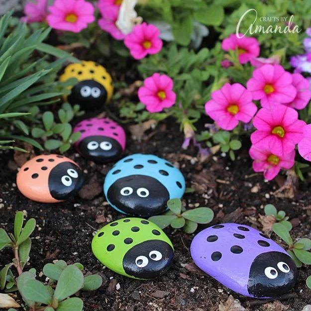 Six smooth rocks painted as ladybugs in yellow, pink, coral, blue, green and purple among pink flowers in a garden