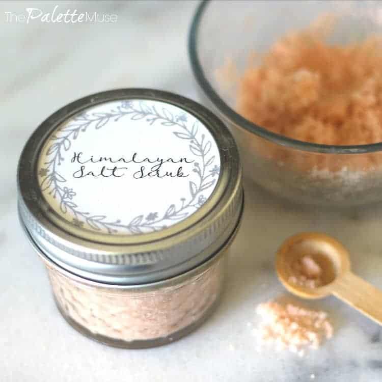 Small mason jar of pink Himalayan salt scrub with a printed lid label beside a bowl of scrub and a wooden measuring spoon