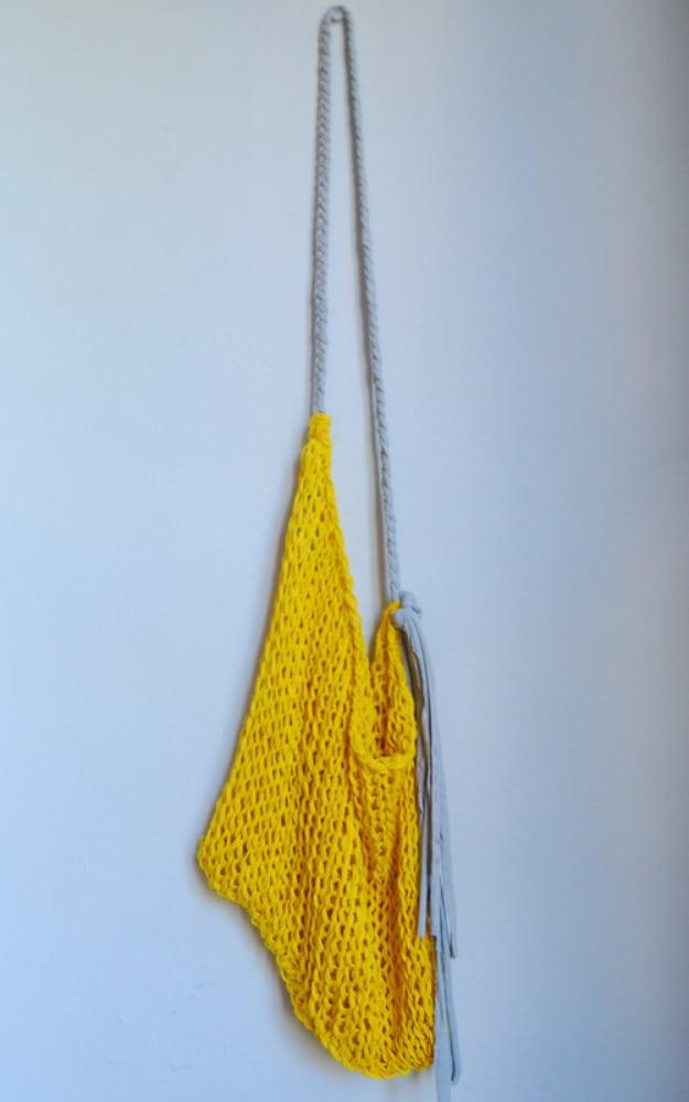 Bright yellow open mesh knitted string bag hanging flat on a pale wall from a long braided fabric strap