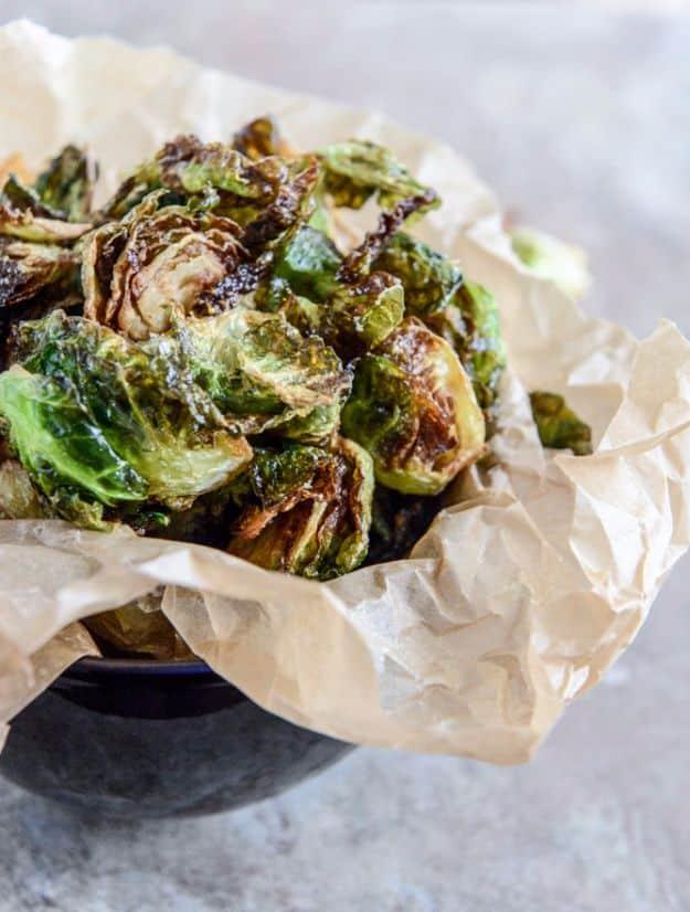 Fried brussels sprout leaves blistered dark and crisp, heaped up in parchment inside a dark metal bowl