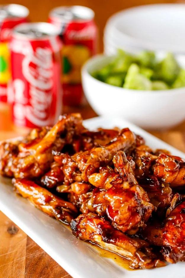 Coca Cola baked chicken wings piled dark and sticky on a white platter with Coke cans and a bowl of chopped celery