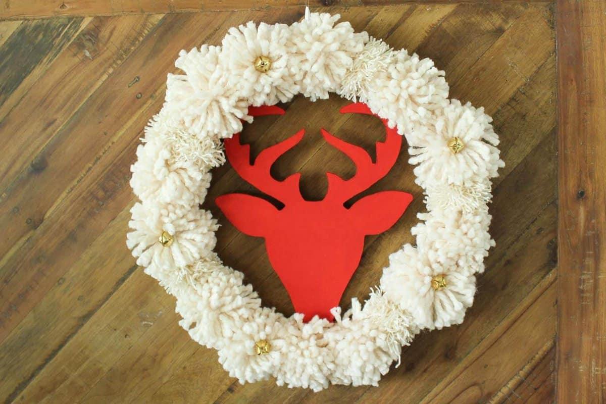 A wreath of large cream yarn pom poms with small gold jingle bells and a red wooden deer head silhouette in the center