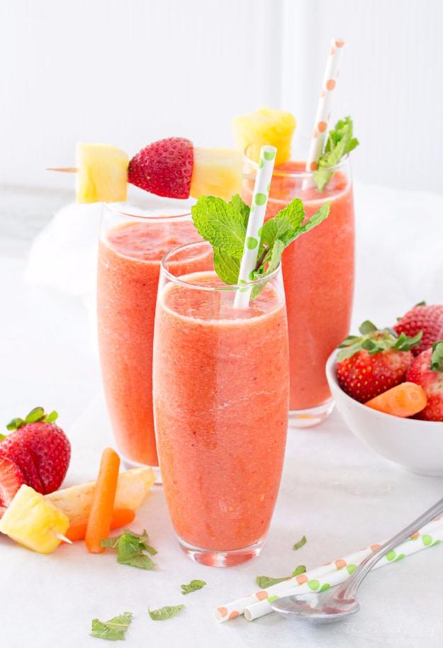Three tall flutes of pink orange tropical carrot smoothie topped with fruit skewers and mint on a white cloth