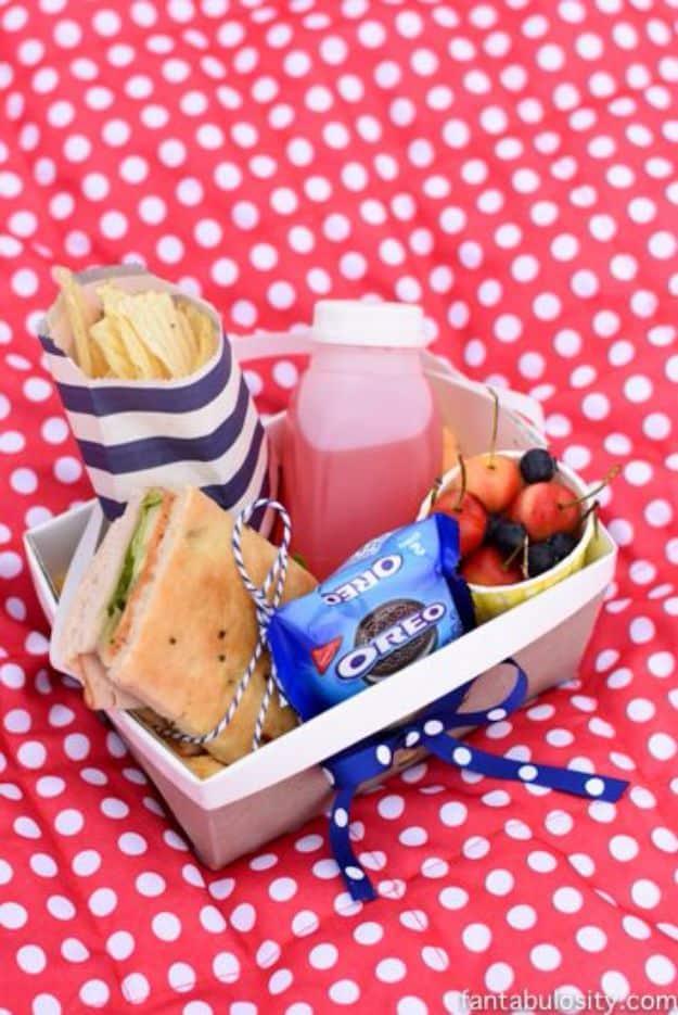 DIY picnic boxed lunch, a white box on a red polka dot cloth holding a wrapped sandwich, chips, fruit and a drink