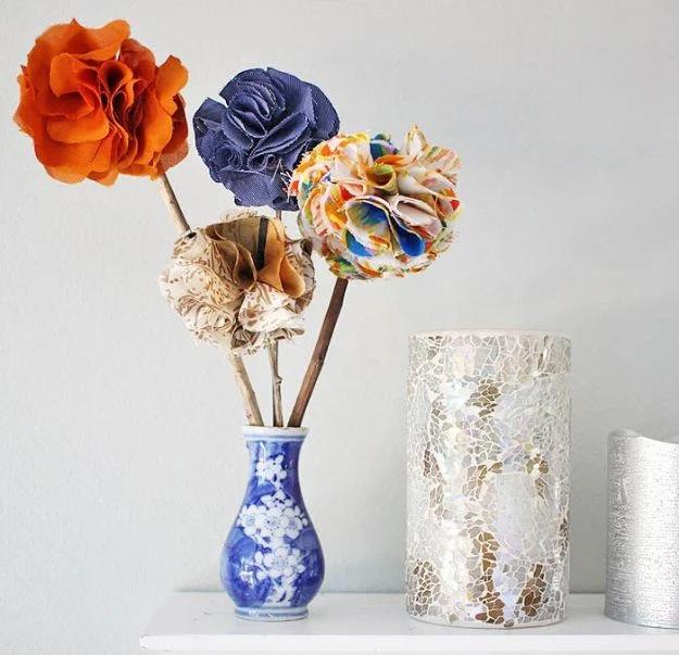 Three handmade fabric flowers in orange, blue and gold on stems in a blue and white vase by a mercury jar