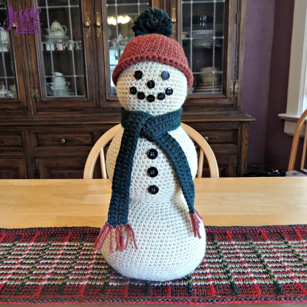 A crocheted cream snowman with button eyes, a rust knit hat with a pom pom and a dark green scarf on a plaid runner